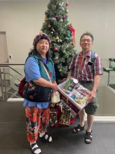 A smiling couple holding a Christmas gift hamper, standing in front of a decorated Christmas tree.