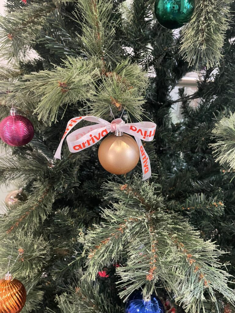 A close-up of the Christmas tree branch with baubles and a gold ornament tied with a ribbon printed with “arriva.”