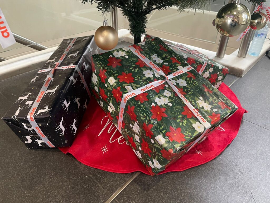 Wrapped Christmas presents under the tree, including a large gift in green and red poinsettia wrapping paper tied with an “arriva” ribbon.