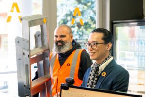 Two arriva team members smiling, one wearing an orange safety vest and the other in a suit with an arriva pin, standing indoors near a ladder.