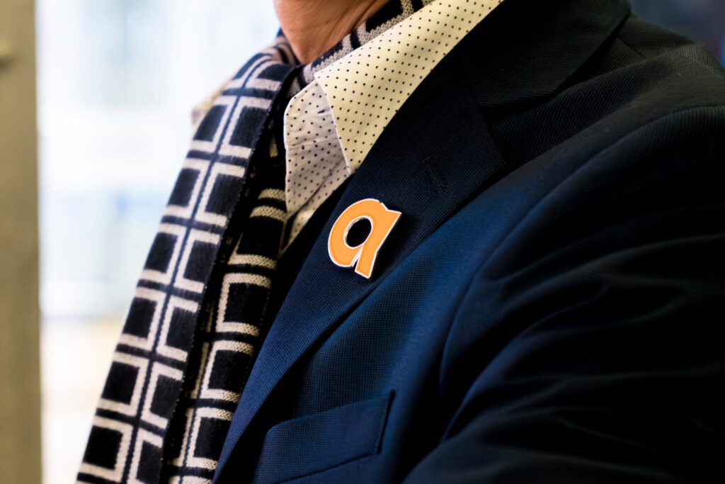 Close-up of a person wearing a suit jacket with an orange arriva “a” pin on the lapel.