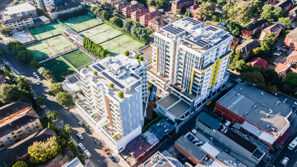 Aerial view of modern residential buildings featuring rooftop solar panels and landscaped terraces, surrounded by city streets and green sports fields. The high-rise towers, possibly Arriva’s Morwick and Lyons buildings in Strathfield, reflect sustainable urban living with energy-efficient design and contemporary architecture.