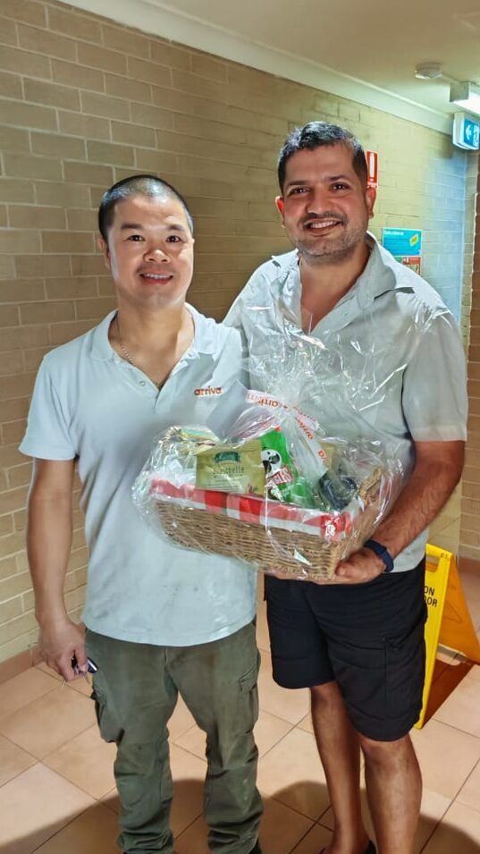 Two men standing together in a hallway, holding a festive basket wrapped in cellophane.