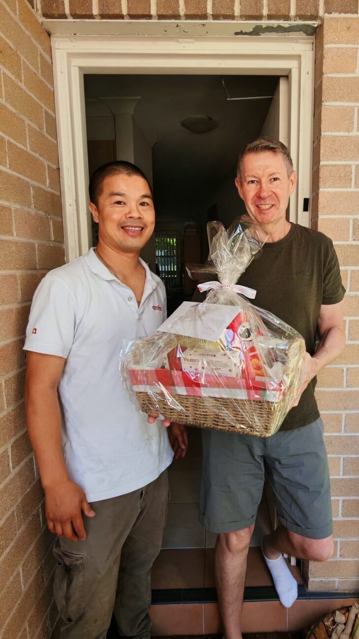 Two men standing at a doorway, one holding a large Christmas gift hamper wrapped in cellophane.