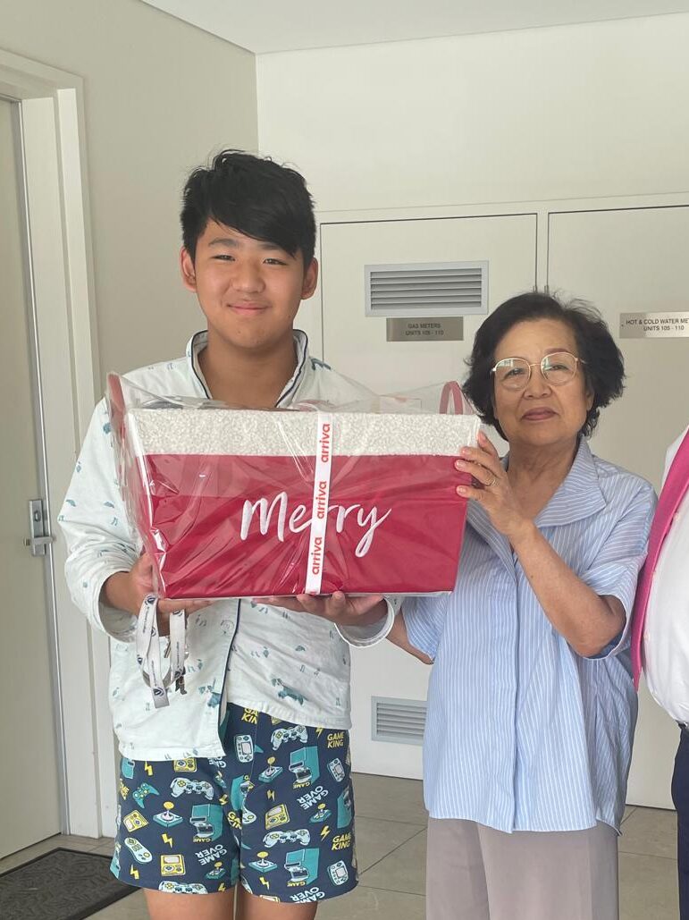 A young man and an older woman smiling while holding a red gift box that says “Merry.”